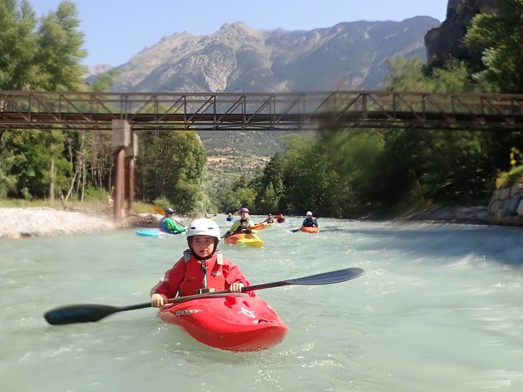 A group of people in kayaks on a river under a bridge
Description automatically generated